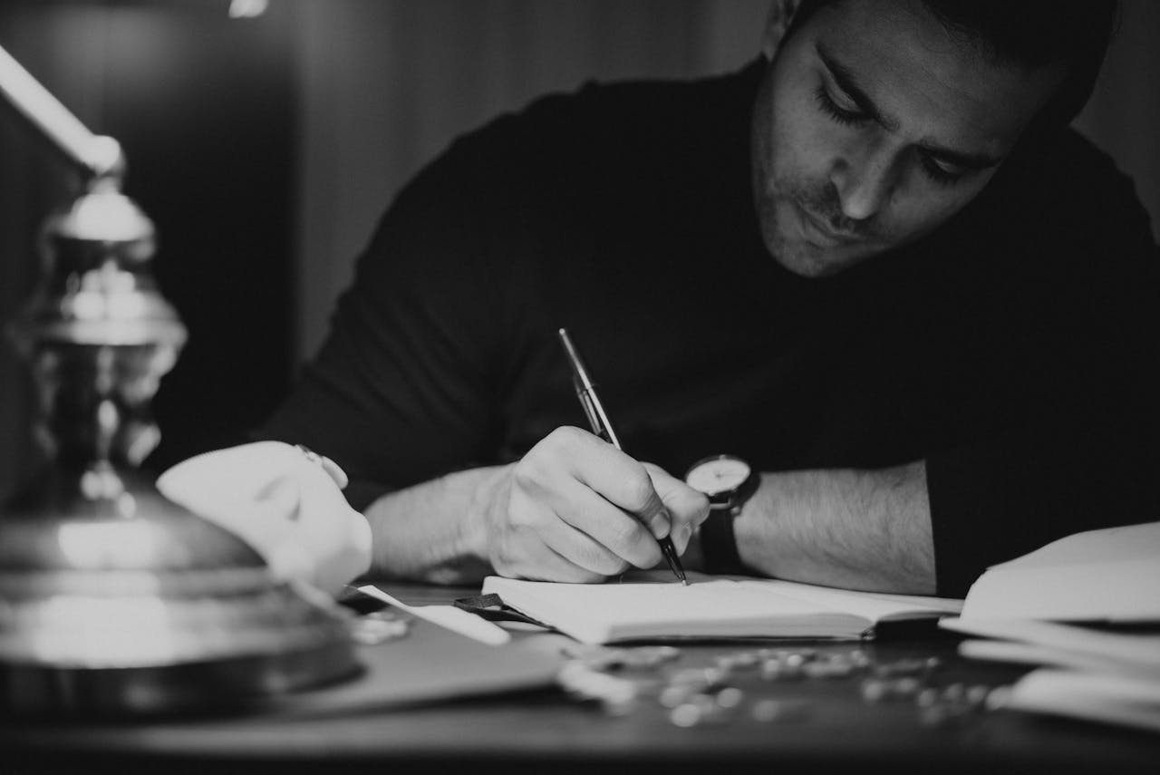 A man writing at a desk under dim lighting, concentrating with intent focus.