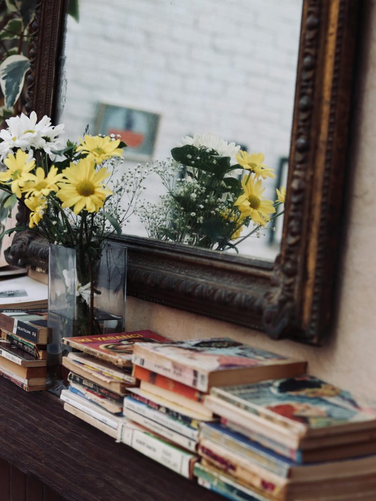 Stack of various books and glass vase with flowers placed on wooden table near mirror in daylight