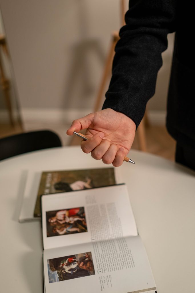 pexels photo 6857707 6857707 A person holding a pen over an open book on a table, symbolizing focus and creativity in a workplace.